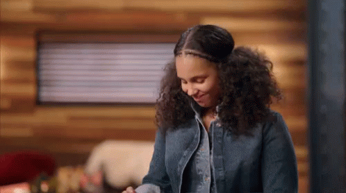 A Woman With Curly Hair Is Standing In Front Of A Wooden Wall In A Cabin And Smiling . GIF