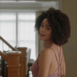 A Woman With Curly Hair Is Standing Next To A Staircase GIF