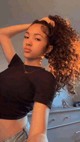 A Woman With Curly Hair Is Wearing A Black Crop Top And Denim Shorts And Holding Her Hair In A Ponytail . GIF