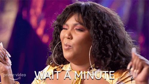 A Woman With Curly Hair Is Wearing A Gold Shirt And Earrings And Says Wait A Minute GIF