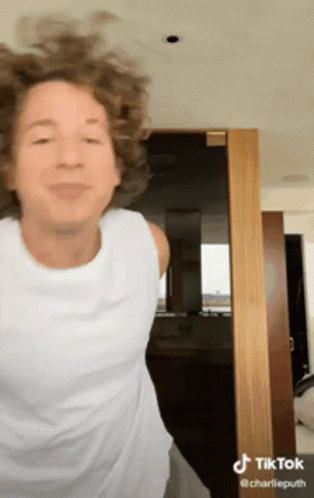 A Woman With Curly Hair Is Wearing A White Tank Top And Dancing In Front Of A Door . GIF