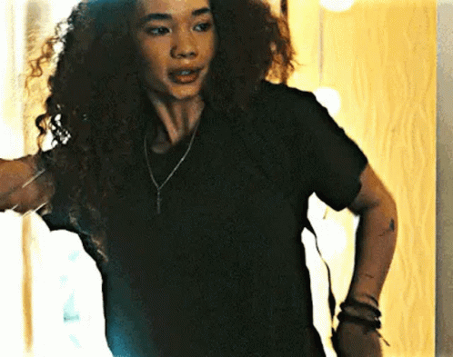 a woman with curly hair wearing a black t-shirt and a necklace is standing in front of a mirror .