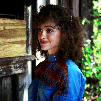 A Woman With Curly Hair Wearing A Blue Jacket Stands In Front Of A Door GIF