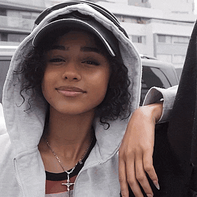 A Woman With Curly Hair Wearing A Hat And A Necklace With A Cross On It GIF
