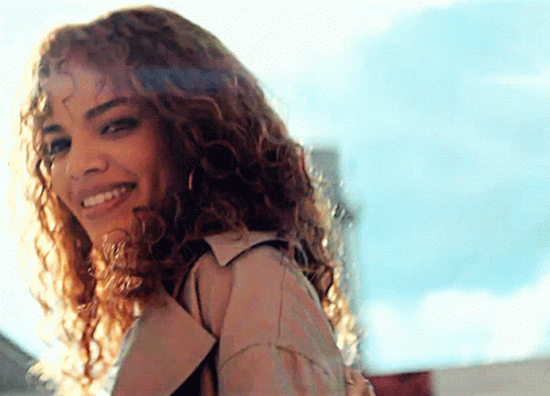 A Woman With Curly Hair Wearing A Trench Coat Is Smiling GIF