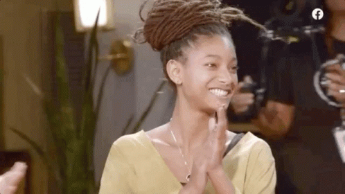 A Woman With Dreadlocks Is Smiling And Clapping Her Hands While Wearing A Yellow Shirt . GIF