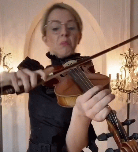 A Woman With Glasses Is Playing A Violin In Front Of A Chandelier GIF