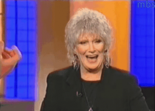 A Woman With Gray Hair Is Smiling In Front Of A Blue Wall With Mbs Written On It GIF