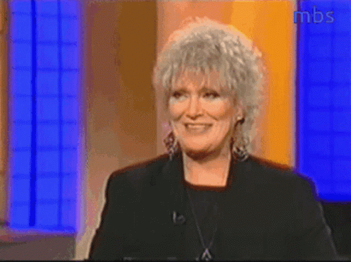 A Woman With Gray Hair Is Smiling In Front Of A Mbs Sign GIF