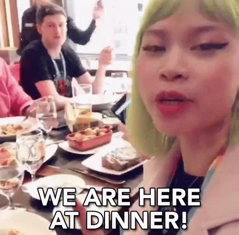 A Woman With Green Hair Is Sitting At A Table With Plates Of Food And Glasses Of Wine And Says We Are Here At Dinner . GIF