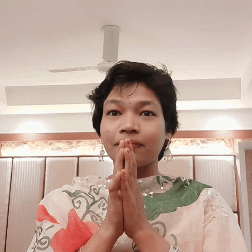 A Woman With Her Hands Folded In Front Of Her Face And A Ceiling Fan In The Background GIF