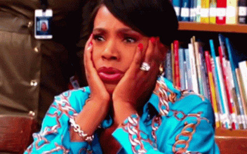 A Woman With Her Hands On Her Face Is Sitting In Front Of A Bookshelf . GIF