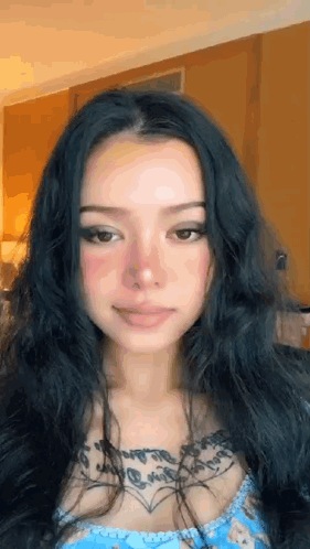 A Woman With Long Black Hair And A Tattoo On Her Chest Is Smiling . GIF