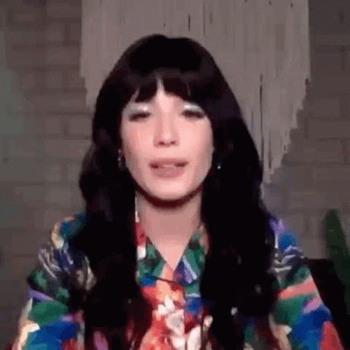 A Woman With Long Black Hair And Bangs Is Wearing A Colorful Shirt And Tie . GIF