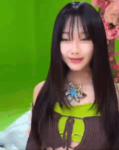 A Woman With Long Black Hair And Bangs Is Wearing A Green Top And A Brown Top . GIF