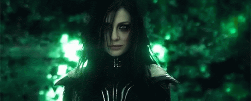 A Woman With Long Black Hair And Green Eyes Is Standing In Front Of A Forest . GIF