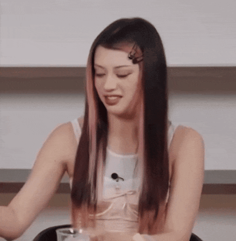 A Woman With Long Black Hair And Pink Streaks Is Sitting At A Table With A Glass Of Water . GIF