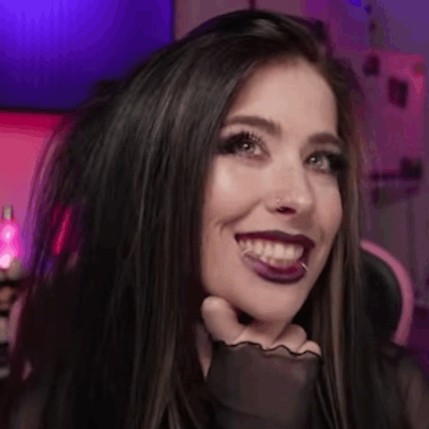 A Woman With Long Black Hair And Purple Lipstick Is Smiling And Holding Her Hand To Her Face . GIF