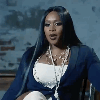 A Woman With Long Black Hair Is Sitting In A Chair Wearing A Blue Jacket And Pearls . GIF