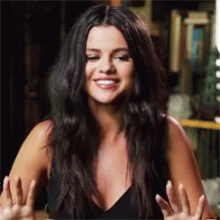 A Woman With Long Black Hair Is Smiling And Making A Funny Face While Sitting At A Table . GIF