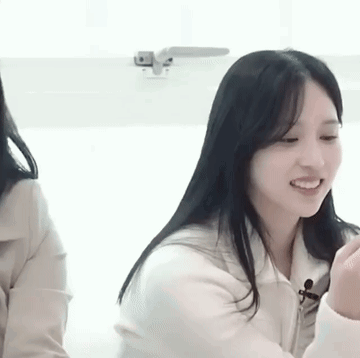 A Woman With Long Black Hair Is Smiling While Sitting Next To Another Woman In A Room . GIF