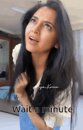A Woman With Long Black Hair Is Standing In A Room With The Words Wait A Minute Below Her . GIF