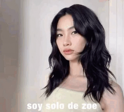 A Woman With Long Black Hair Is Standing In Front Of A Wall And Looking At The Camera . GIF
