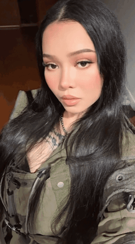 A Woman With Long Black Hair Is Wearing A Camouflage Jacket And Necklace GIF