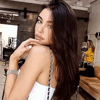 A Woman With Long Black Hair Is Wearing A White Tank Top With A Chain Strap . GIF