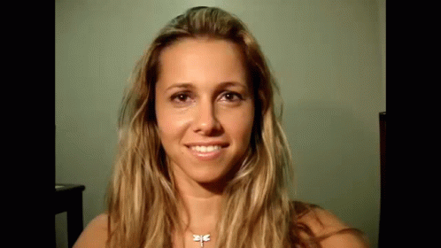 A Woman With Long Blonde Hair And A Dragonfly Necklace Is Smiling . GIF