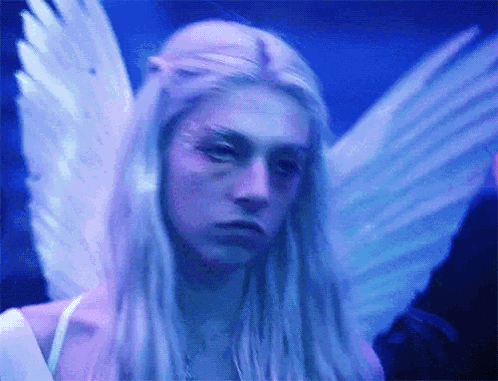A Woman With Long Blonde Hair And Angel Wings Is Looking At The Camera In A Dark Room . GIF