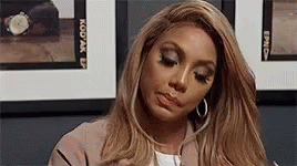 A Woman With Long Blonde Hair And Hoop Earrings Is Sitting In Front Of A Wall Of Pictures . GIF