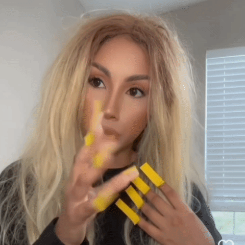 A Woman With Long Blonde Hair Is Applying Yellow Nail Polish To Her Nails GIF