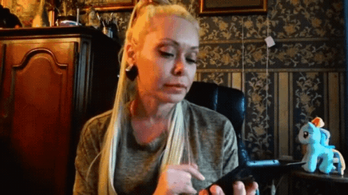 A Woman With Long Blonde Hair Is Sitting In A Chair Looking At Her Phone . GIF