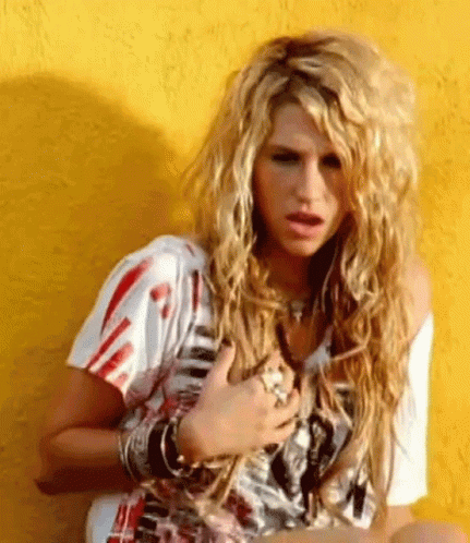 A Woman With Long Blonde Hair Is Sitting In Front Of A Yellow Wall With Her Hands On Her Chest . GIF