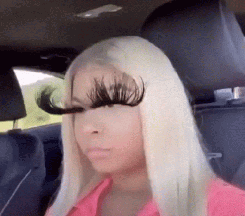 A Woman With Long Blonde Hair Is Sitting In The Back Seat Of A Car With A Pair Of Fake Eyelashes On Her Face . GIF