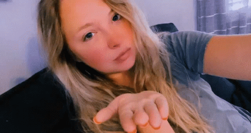 A Woman With Long Blonde Hair Is Sitting On A Couch Taking A Selfie . GIF