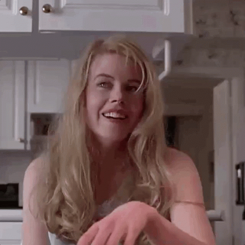 A Woman With Long Blonde Hair Is Smiling In A Kitchen While Holding Her Hands Together . GIF