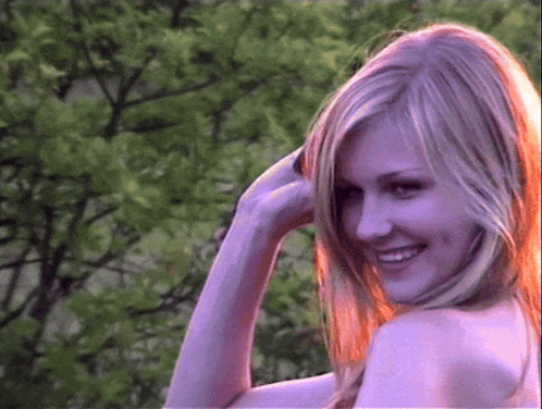 A Woman With Long Blonde Hair Is Smiling In Front Of A Tree GIF