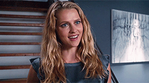 A Woman With Long Blonde Hair Is Smiling In Front Of Stairs And A Painting GIF