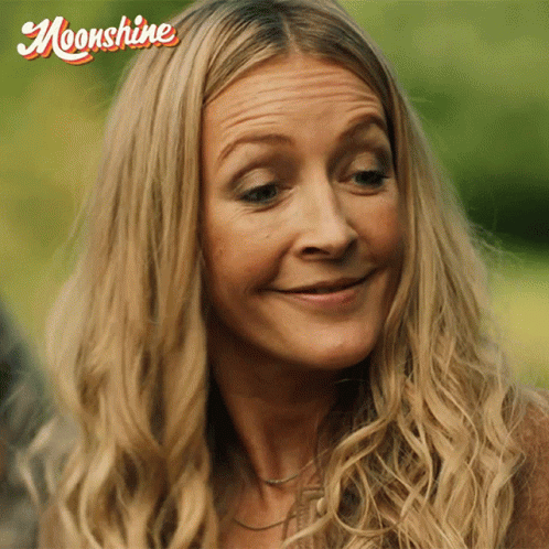 A Woman With Long Blonde Hair Is Smiling With A Moonshine Logo Behind Her GIF