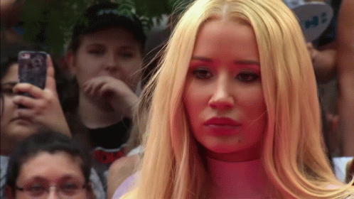 A Woman With Long Blonde Hair Is Standing In Front Of A Crowd While A Man Takes A Picture With His Phone GIF