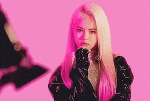 A Woman With Long Blonde Hair Is Standing In Front Of A Pink Background With Her Hands On Her Chin . GIF