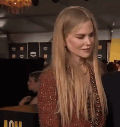 A Woman With Long Blonde Hair Is Standing In Front Of A Table And Looking At Something . GIF
