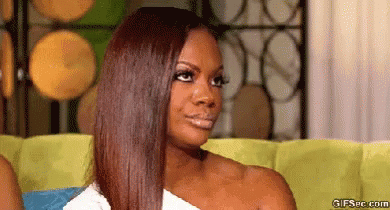 A Woman With Long Brown Hair Is Sitting On A Couch And Making A Funny Face . GIF