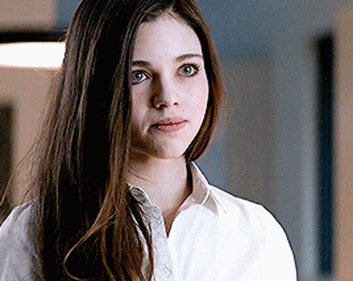 A Woman With Long Brown Hair Is Wearing A White Shirt And Looking At The Camera . GIF