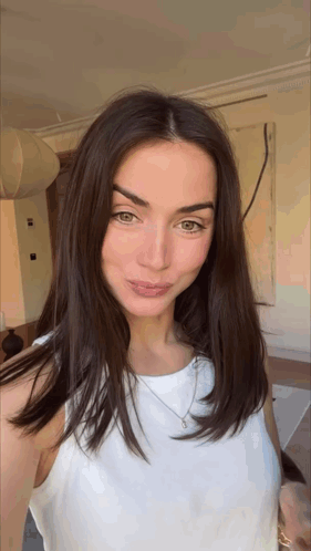 a woman with long brown hair is wearing a white tank top and necklace .