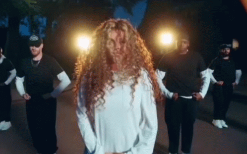 A Woman With Long Curly Hair Is Standing In Front Of A Group Of Men Dancing . GIF