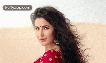A Woman With Long Curly Hair Is Wearing A Red Saree And Earrings And Looking At The Camera . GIF