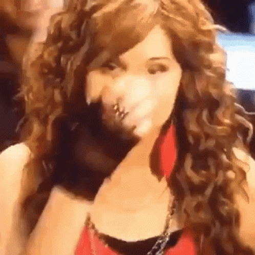 A Woman With Long Curly Hair Is Wearing A Red Top And Earrings And Covering Her Face With Her Hand . GIF